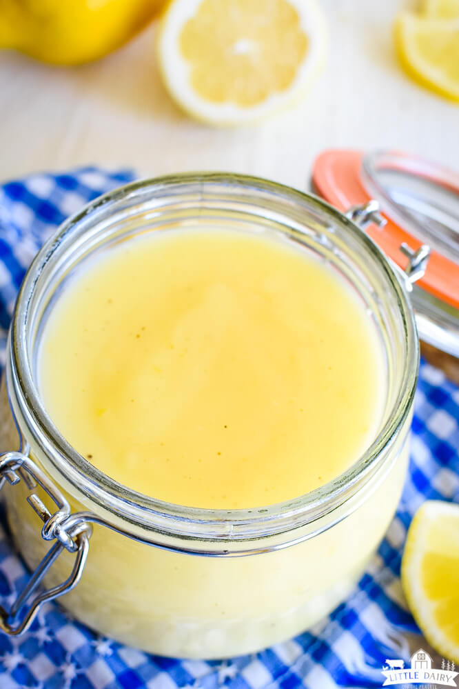 een close up shot van lichtgele lemon curd in een glazen pot boven een blauw-wit geblokt servet. Little Dairy on the Prairie