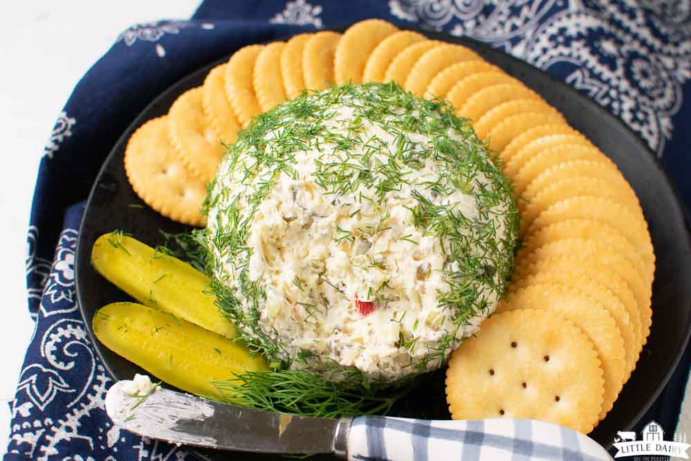 A black plate with a cheese ball covered with fresh dill weed and a knife for spreading cream cheese dip. Surrounded by Ritz crackers. Little Dairy on the Prairie
